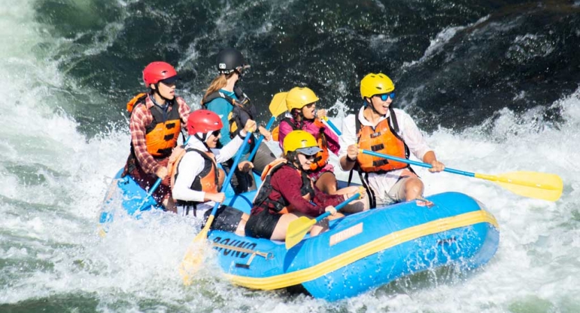 a group of outward bound students wearing helmets and life jackets paddle a raft through whitewater